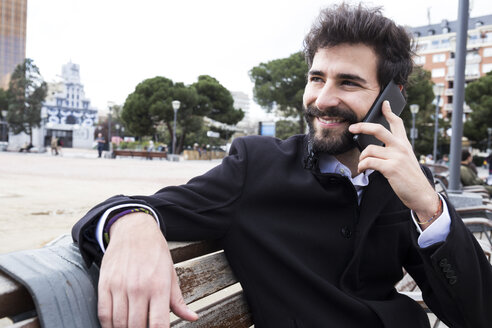 Porträt eines lächelnden jungen Mannes mit Vollbart, der auf einer Bank sitzend telefoniert - ABZF01982