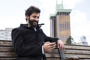 Spanien, Madrid, lächelnder junger Mann mit Vollbart sitzt auf einer Bank und schaut auf sein Handy - ABZF01984
