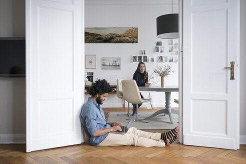 Mann zu Hause auf dem Boden sitzend, mit Laptop arbeitend, mit Frau im Hintergrund - SBOF00381