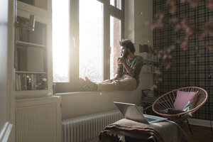 Mann sitzt auf der Fensterbank im Wohnzimmer und schaut mit einer Tasse nach draußen - SBOF00388
