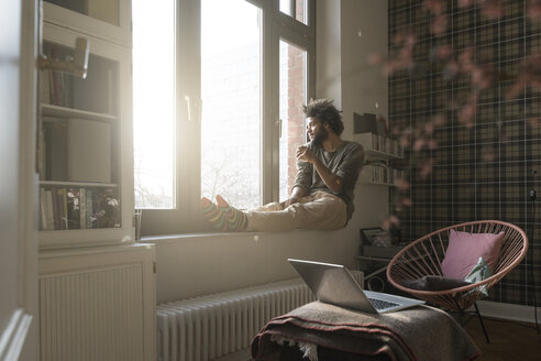 Man sitting on window sill in living room looking outside holding a cup - SBOF00388