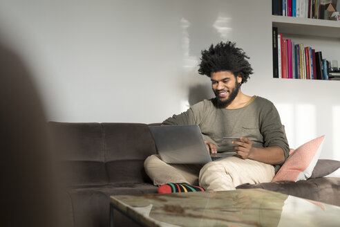 Mann sitzt auf der Couch im Wohnzimmer mit Laptop und Kreditkarte - SBOF00393
