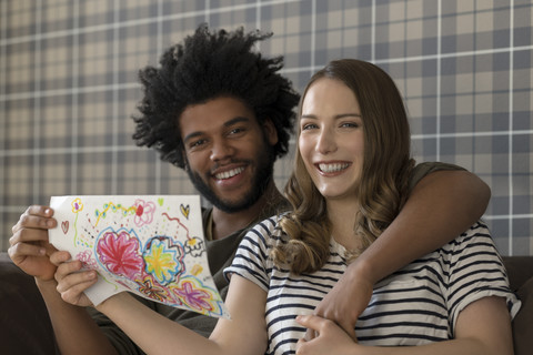 Lächelndes Paar sitzt auf der Couch und zeigt eine Kinderzeichnung, lizenzfreies Stockfoto