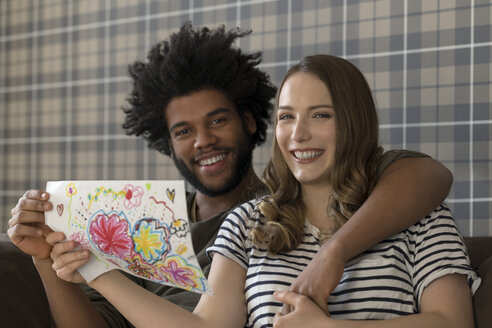 Lächelndes Paar sitzt auf der Couch und zeigt eine Kinderzeichnung - SBOF00400