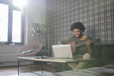 Man sitting in living room on sofa playing guitar in front of laptop - SBOF00402