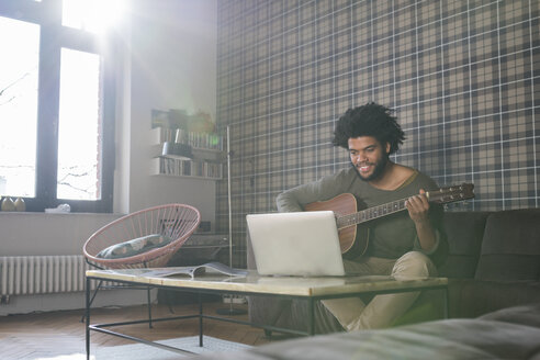 Mann sitzt im Wohnzimmer auf dem Sofa und spielt Gitarre vor einem Laptop - SBOF00402