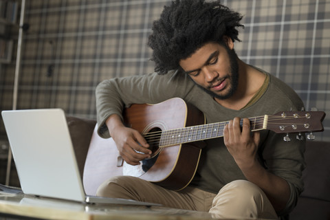 Mann sitzt im Wohnzimmer auf dem Sofa und spielt Gitarre vor einem Laptop, lizenzfreies Stockfoto