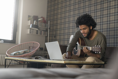 Mann sitzt im Wohnzimmer auf dem Sofa und spielt Gitarre vor einem Laptop - SBOF00404