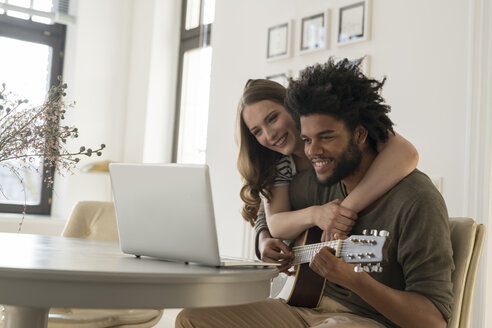 Lächelndes Paar vor einem Laptop im Wohnzimmer, Mann spielt Gitarre - SBOF00408