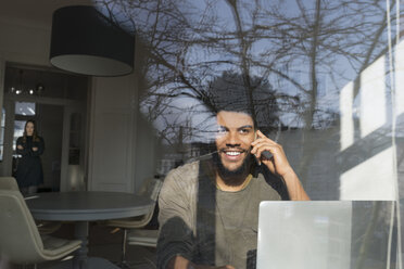 Man speaking on smartphone in front of laptop, woman listening in the back - SBOF00416