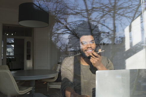 Mann, der vor einem Laptop am Fenster mit seinem Smartphone spricht - SBOF00417