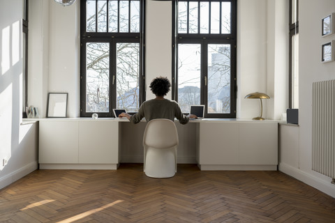 Mann arbeitet vor einem Panoramafenster zu Hause mit Laptop und Tablet, lizenzfreies Stockfoto