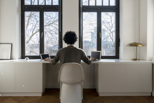 Mann arbeitet vor einem Panoramafenster zu Hause mit Laptop und Tablet - SBOF00420