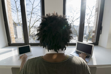 Mann arbeitet vor einem Panoramafenster zu Hause mit Laptop und Tablet - SBOF00421