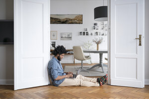 Mann sitzt zu Hause auf dem Boden und arbeitet mit Laptop im Türrahmen - SBOF00444