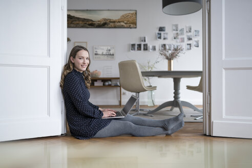 Lächelnde Frau zu Hause, die auf dem Boden sitzt und mit einem Laptop im Türrahmen arbeitet - SBOF00445