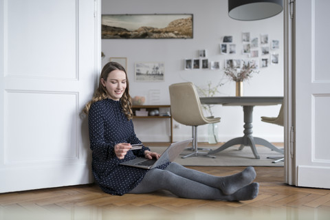 Lächelnde Frau zu Hause auf dem Boden sitzend mit Laptop im Türrahmen, der eine Kreditkarte hält, lizenzfreies Stockfoto