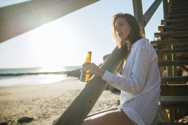 Young woman with bottle of beer relaxing on the beach at sunset - KIJF01464