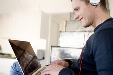 Smiling man using laptop and headphones at home - FMOF00269