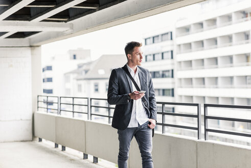 Junger Geschäftsmann, der ein Smartphone benutzt und auf einem Parkdeck spazieren geht - UUF10591