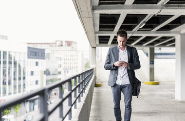 Junger Geschäftsmann, der ein Smartphone benutzt und auf einem Parkdeck spazieren geht - UUF10593