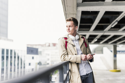 Junger Geschäftsmann mit Smartphone, stehend auf dem Parkdeck - UUF10598