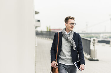 Young businessman walking in the street, carrying digital tablet and ear phones - UUF10604