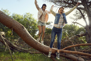 Zwei junge Frauen springen Hand in Hand von einem Baumstamm - SRYF00441