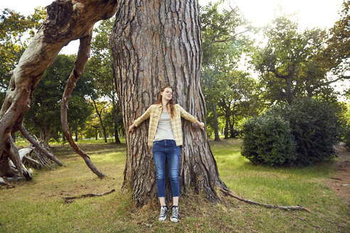 Lächelnde junge Frau lehnt an einem Baumstamm - SRYF00449