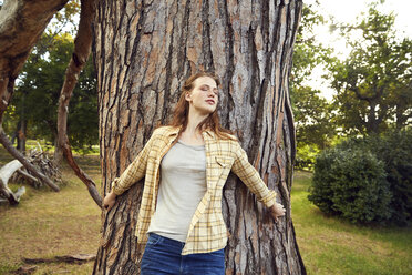 Redheaded young woman leaning against tree trunk with eyes closed - SRYF00451