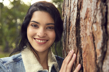 Portrait of smiling young woman beside tree trunk - SRYF00460