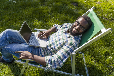 Lächelnder Mann mit Laptop, der auf einem Liegestuhl auf einer Wiese sitzt und zur Kamera aufschaut - TCF05410