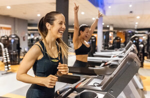 Zwei glückliche Frauen trainieren im Fitnessstudio auf einem Laufband - MGOF03276