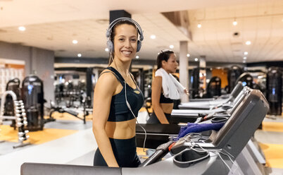 Lächelnde Frau mit Kopfhörern im Fitnessstudio auf einem Laufband - MGOF03278