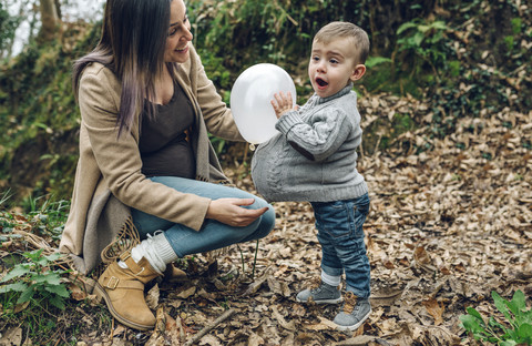 Glückliche schwangere Mutter und kleiner Junge haben Spaß mit einem Luftballon im Wald, lizenzfreies Stockfoto