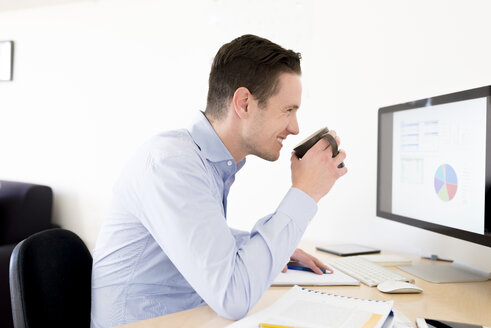 Lächelnder Geschäftsmann mit einer Tasse Kaffee, der ein Tortendiagramm auf einem Computerbildschirm analysiert - FMOF00270