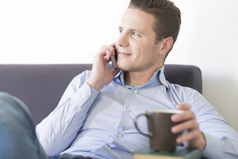 Lächelnder Geschäftsmann auf der Couch mit Smartphone und einer Tasse Kaffee - FMOF00271