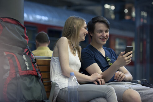 Junges Paar mit Rucksäcken sitzt am Bahnhof und benutzt sein Smartphone - MVCF00167