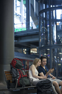 Young couple with backpacks sitting at rail station using smartphone - MVCF00168