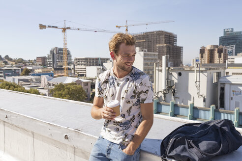 Young man standing on rooftop terrace drinking coffee - WESTF23064