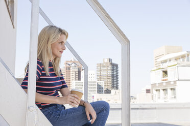 Young woman drinking coffee, sitting on stairs on a rooftop terrace - WESTF23075