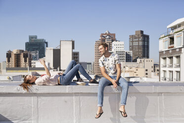 Young couple sitting on balustrade of a rooftop terrace, using smartphone - WESTF23082