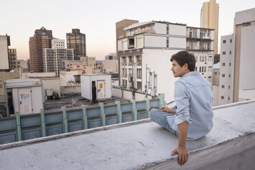 Junger Mann sitzt auf der Brüstung einer Dachterrasse und betrachtet die Aussicht - WESTF23146