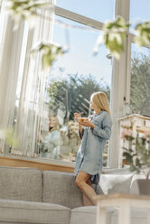Woman at home standing on couch eating fruit salad looking out of window - JOSF00839