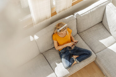 Frau zu Hause auf der Couch sitzend mit Kopfhörern und Tablet - JOSF00867