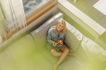 Happy woman at home sitting on couch eating fruit salad - JOSF00876