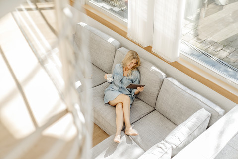 Frau zu Hause auf der Couch mit einer Tasse Kaffee und einem Tablet, lizenzfreies Stockfoto