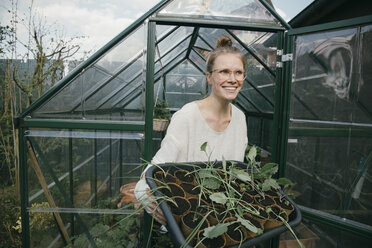 Portrait of happy young woman with potted seedlings in front of greenhouse - MFF03488