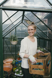 Porträt einer lächelnden jungen Frau mit einer Kiste mit Gartengeräten vor einem Gewächshaus - MFF03489