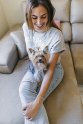 Lächelnde junge Frau sitzt mit ihrem Yorkshire Terrier auf der Couch - KIJF01480